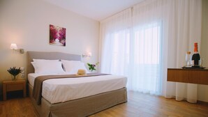 A neatly arranged hotel room with a large bed, a bedside table with a vase of flowers, a wall-mounted lamp, and a window with sheer curtains. - Ionian Heaven Villas (Lefkada)