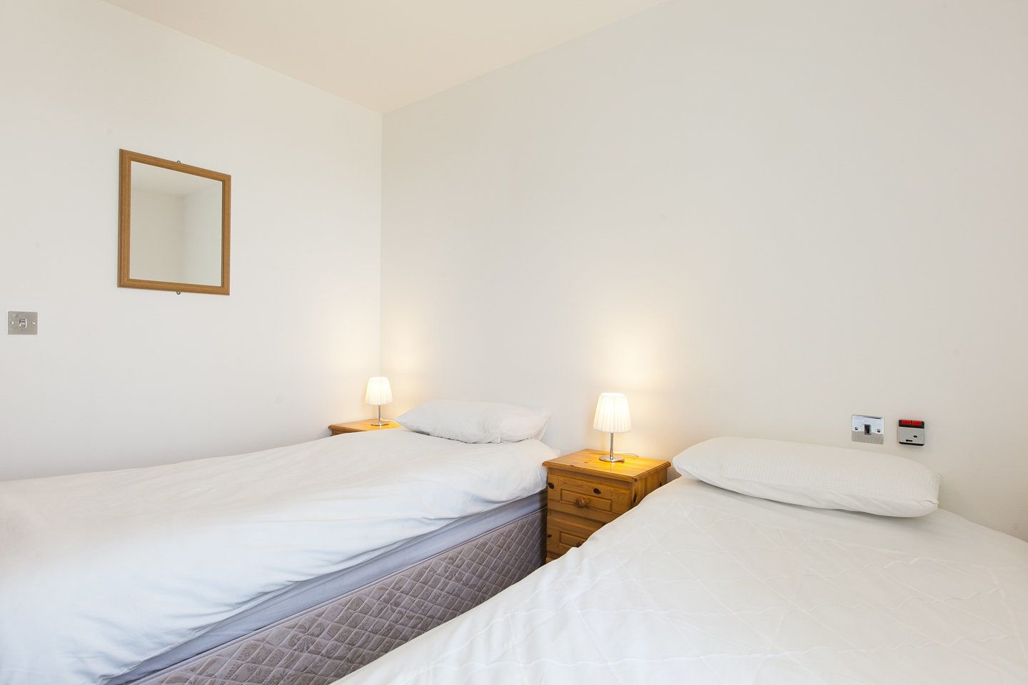 Habitación doble con dos camas individuales, una mesita de noche con lámpara, un espejo en la pared y un termostato.