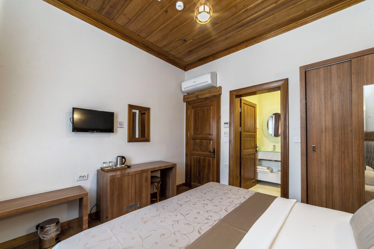 A bedroom with a wooden ceiling, a bed with patterned bedding, a desk with a chair, a TV, and a wardrobe.