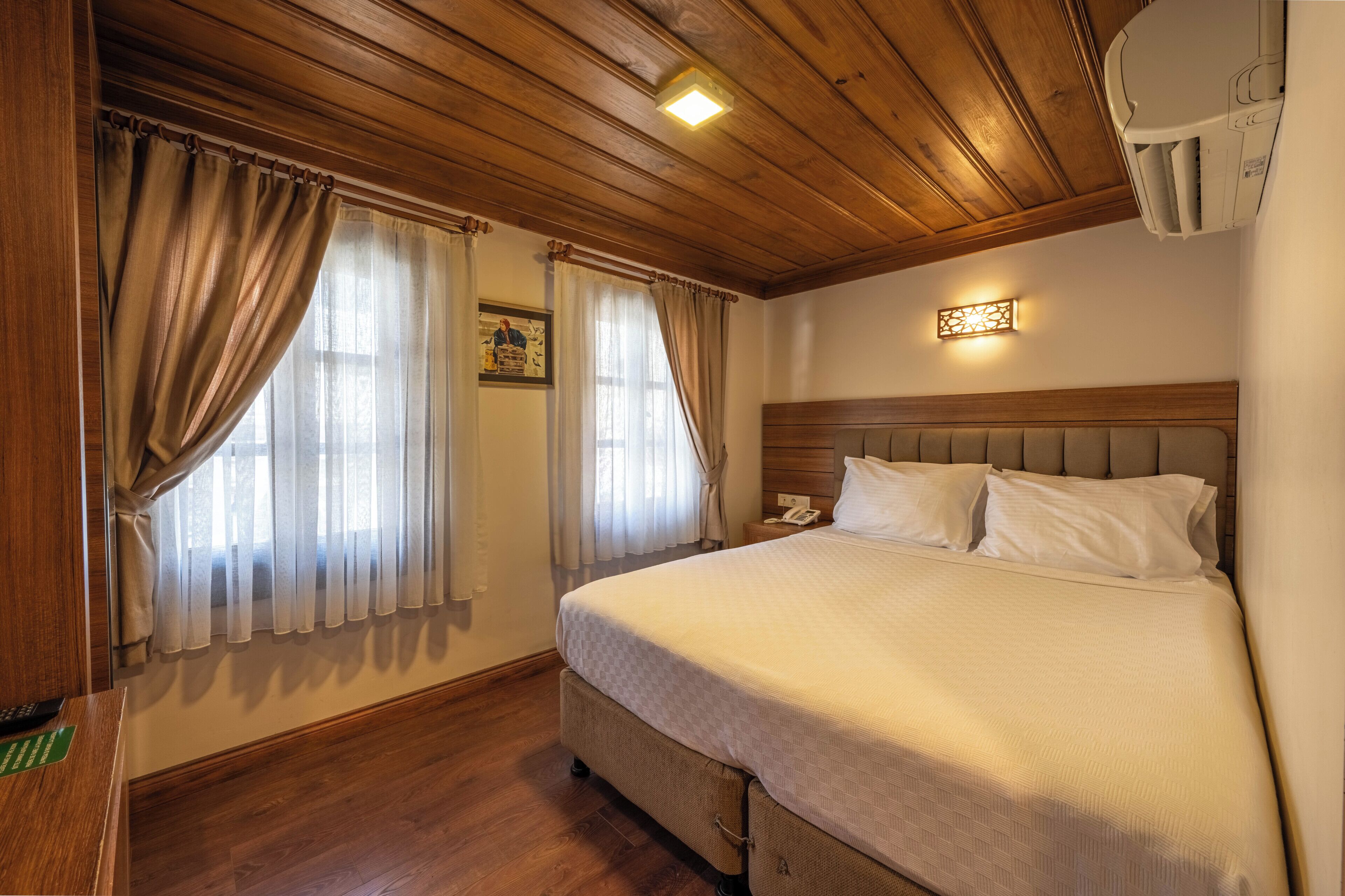 Habitación de hotel con techo de madera, cama con cabecera, mesita de noche con lámpara y ventana con cortinas.