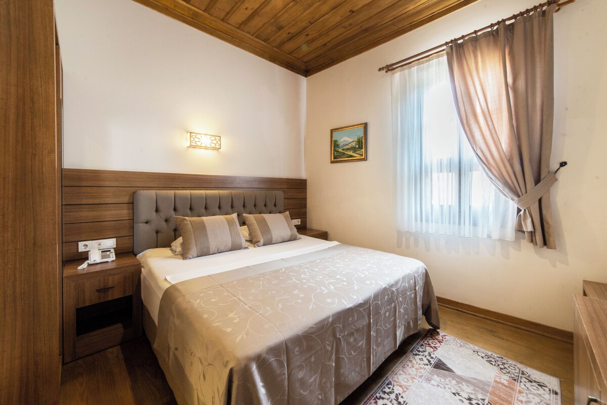 Une chambre à coucher avec une tête de lit en bois, une table de chevet avec un téléphone, un lit recouvert d’une couvre-lit à motifs, un tableau au mur et une fenêtre avec des rideaux.