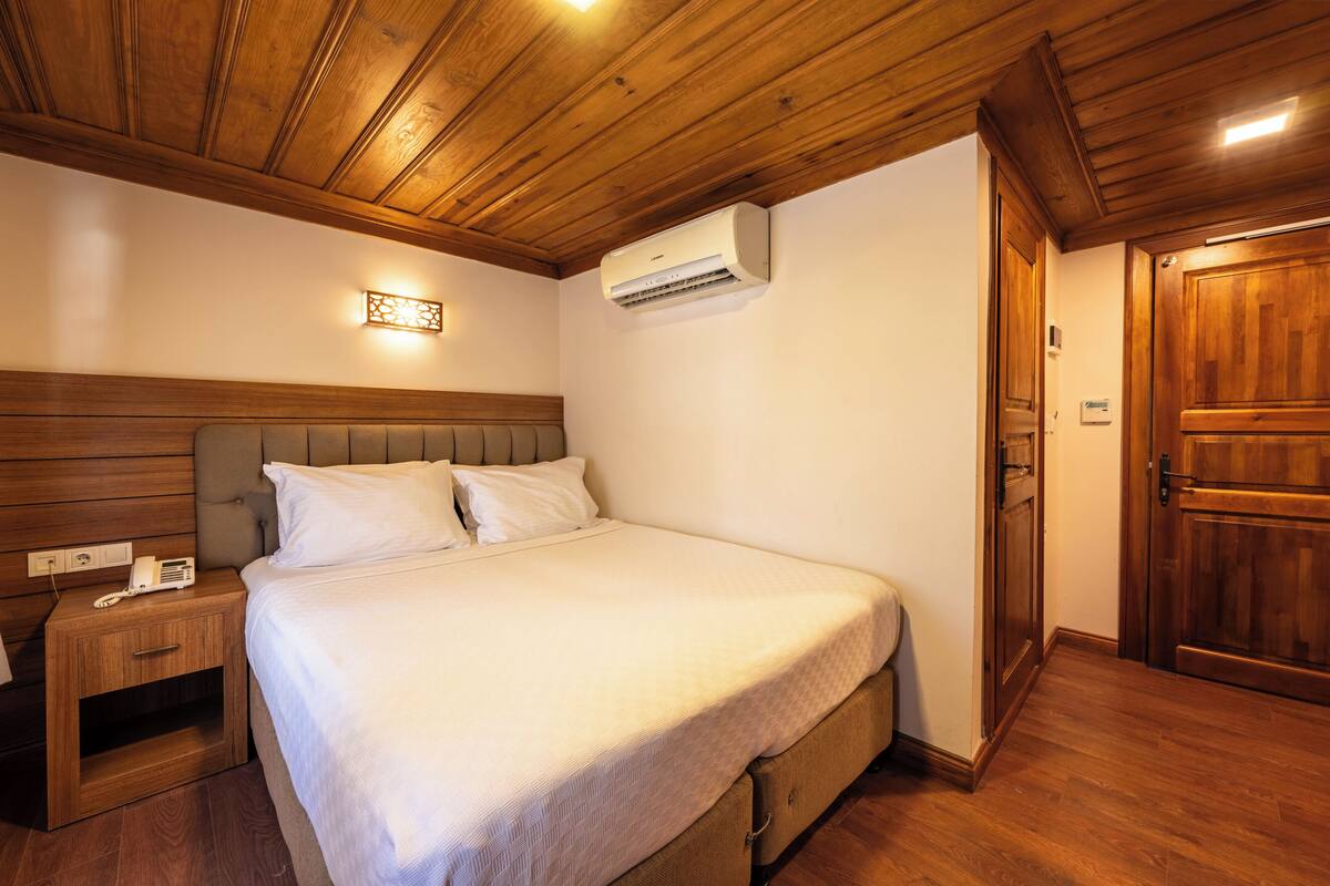 A hotel room with a wooden ceiling, a bed with white bedding, an air conditioning unit, a bedside telephone, and a wooden door.