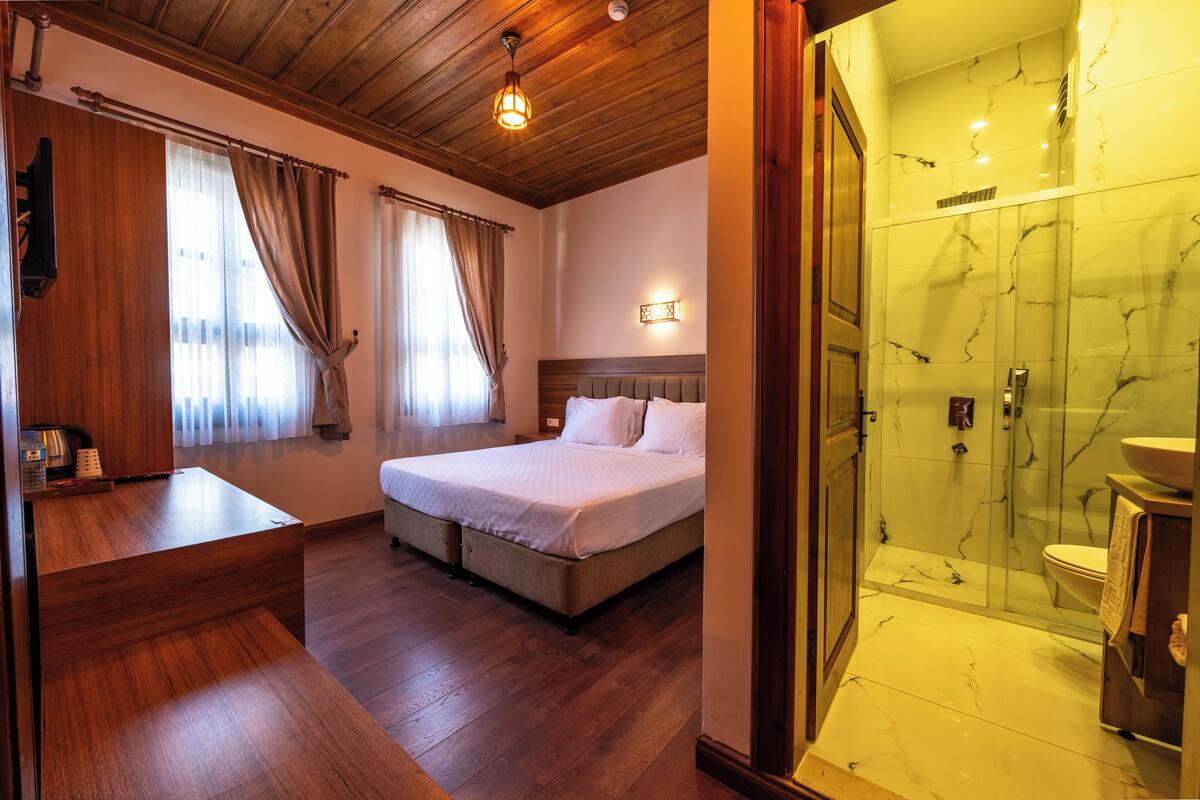 A hotel room with a wooden ceiling, a bed with white bedding, a bedside table, a shower with a glass enclosure, and a marble-tiled wall.