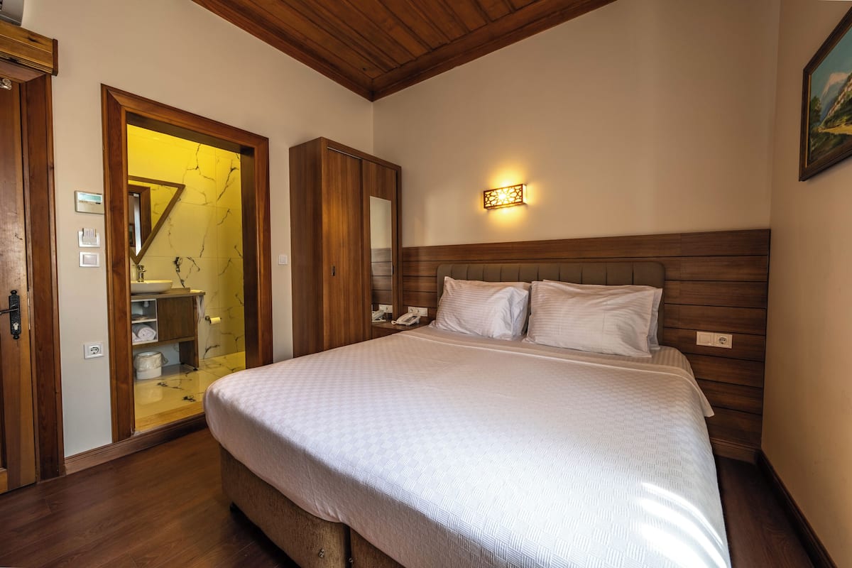 A bedroom with a wooden headboard, a bed with white bedding, a nightstand, a wall-mounted light, and a bathroom visible through an open door.