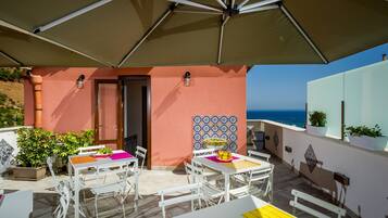 Un patio exterior con mesas y sillas blancas, un paraguas grande y vista al mar.