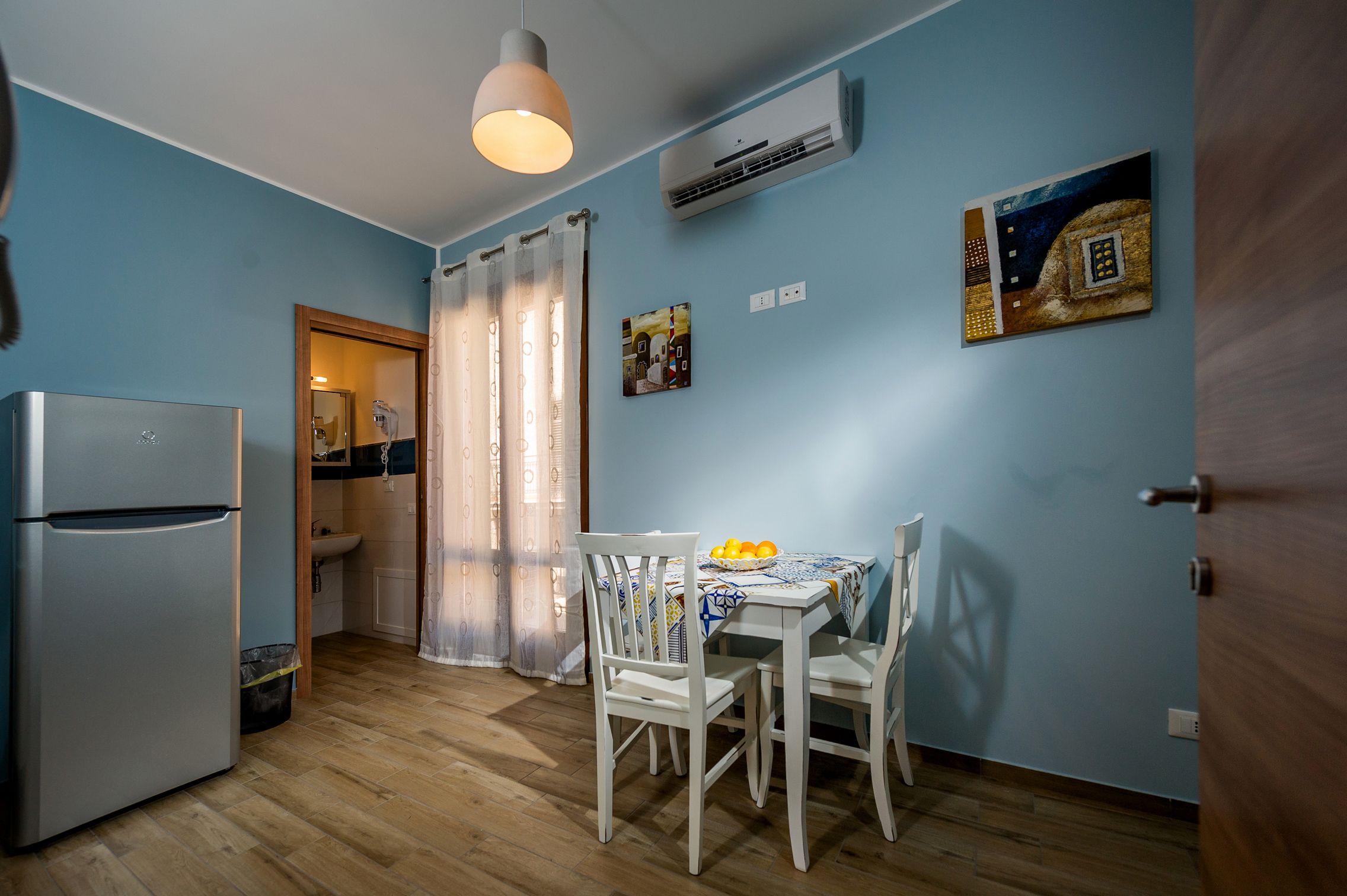 A compact kitchen with a refrigerator, a dining table with chairs, and a hanging pendant light.