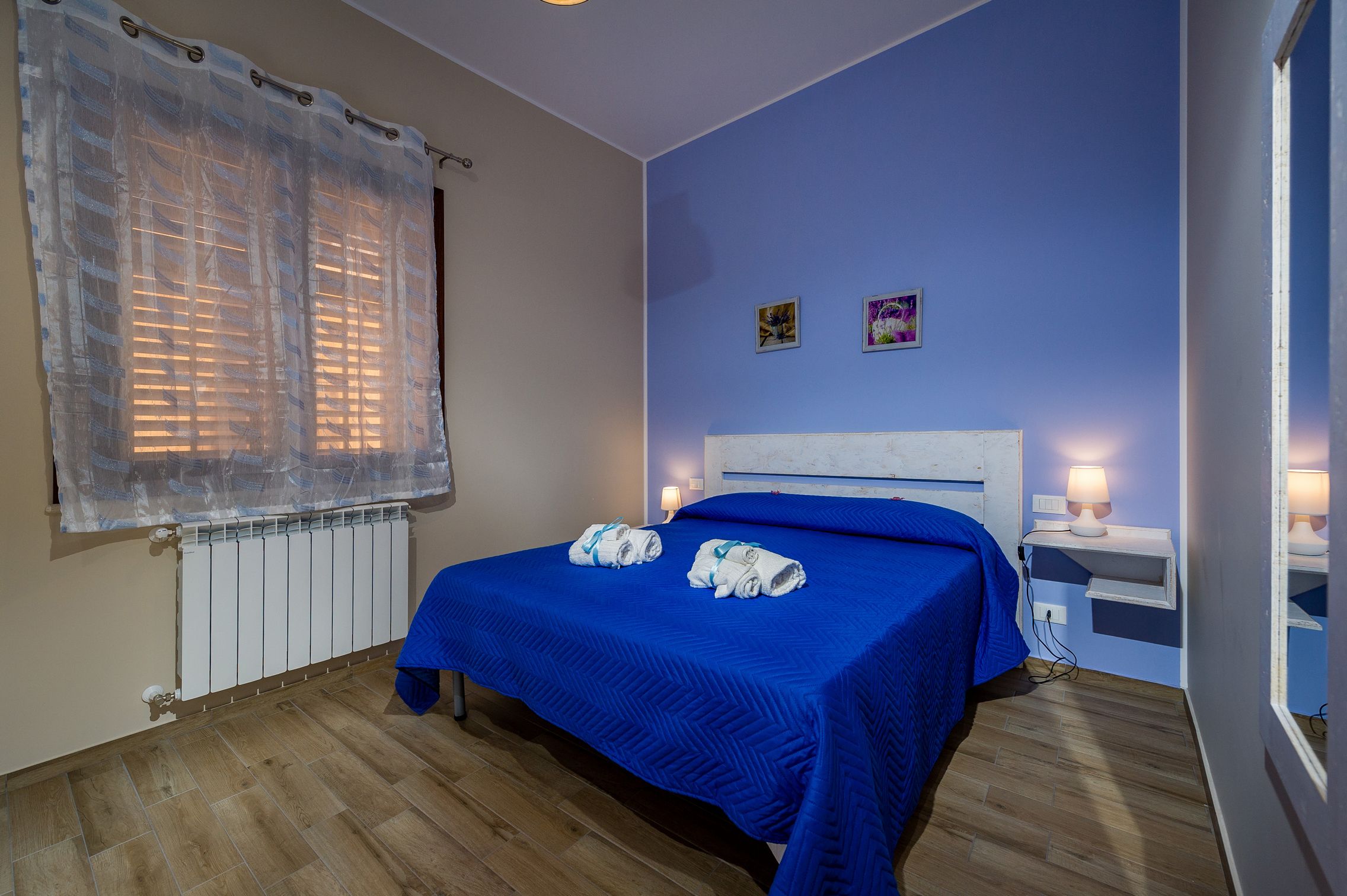A bedroom with a blue bedspread, a white headboard, a bedside table with a lamp, and a window with blinds.