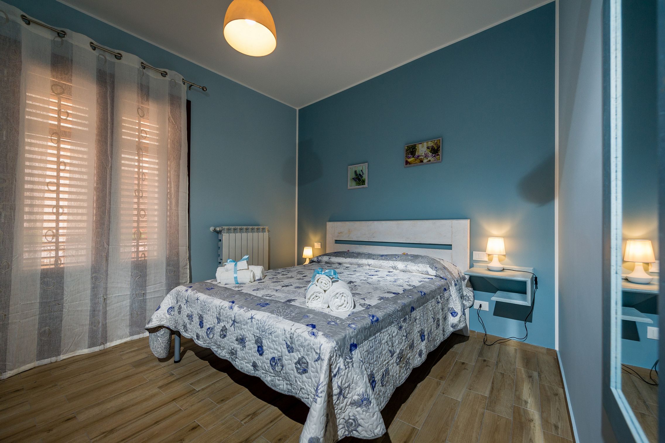 A bedroom with a bed, a bedside table with a lamp, a radiator, and a window with curtains.