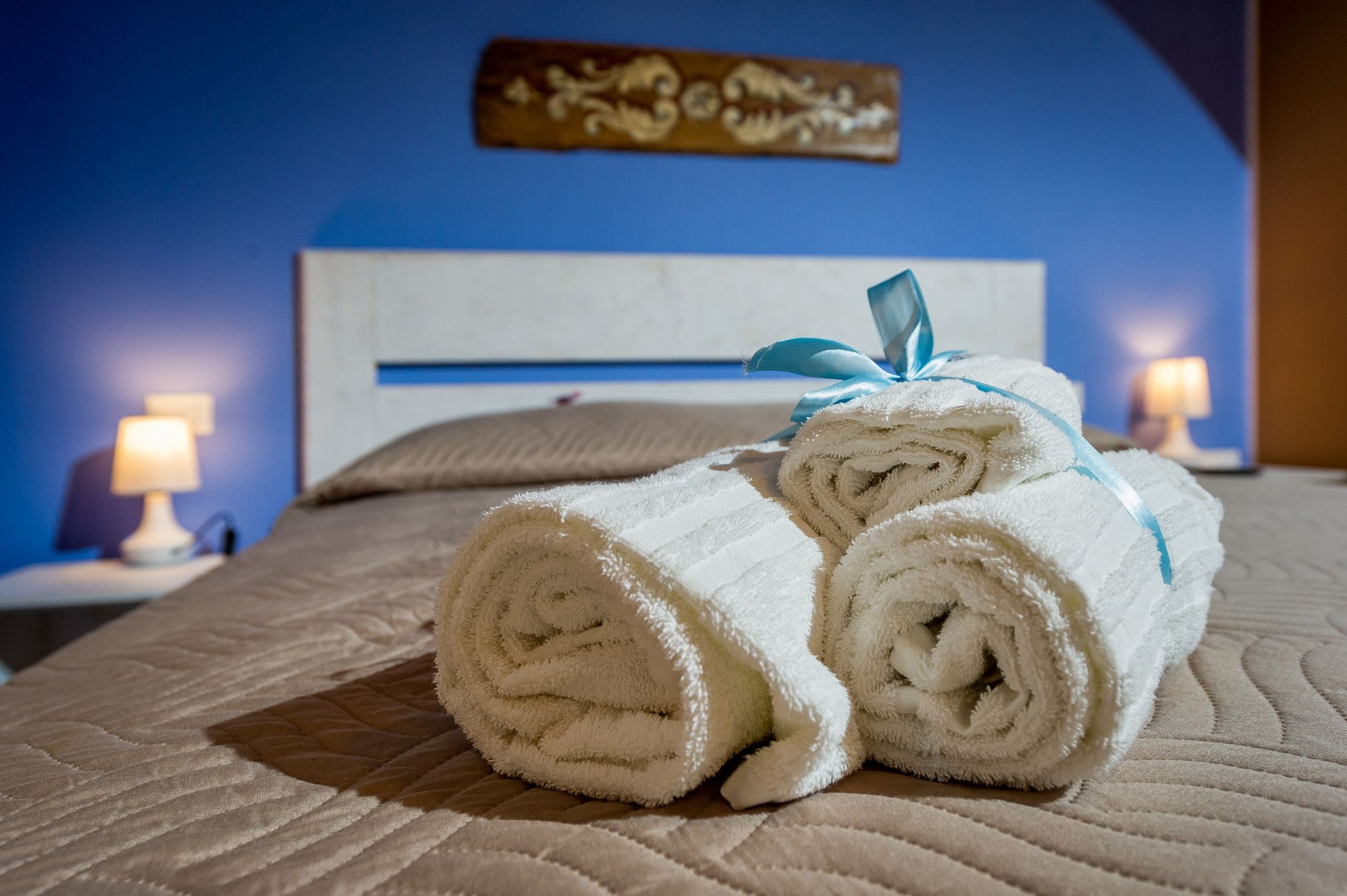 A neatly made bed with rolled white towels tied with blue ribbons, a blue wall, and a decorative piece above the bed.