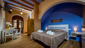 Quarto de hotel com cama, escrivaninha com cadeiras, televisão e parede azul de destaque.