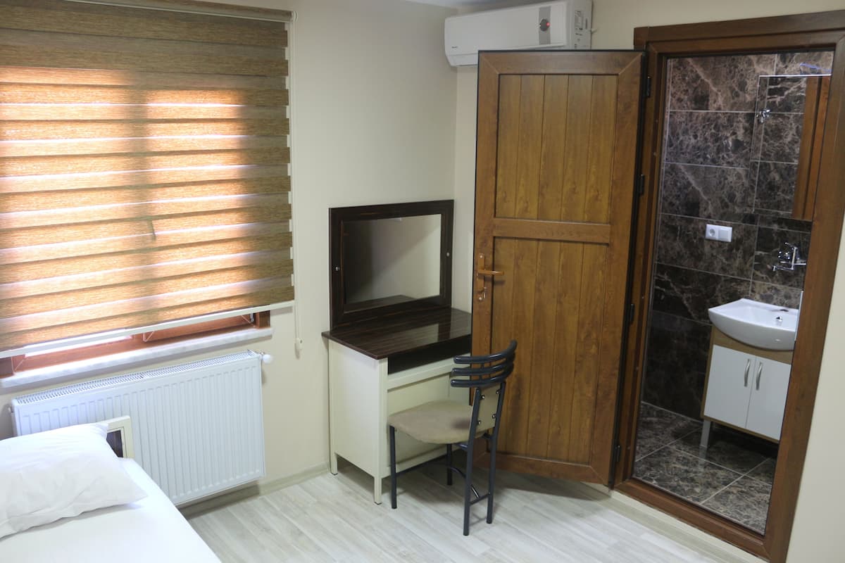 A bedroom with a bed, a desk with a chair, a wooden wardrobe, a bathroom with a sink and mirror, and a window with blinds.