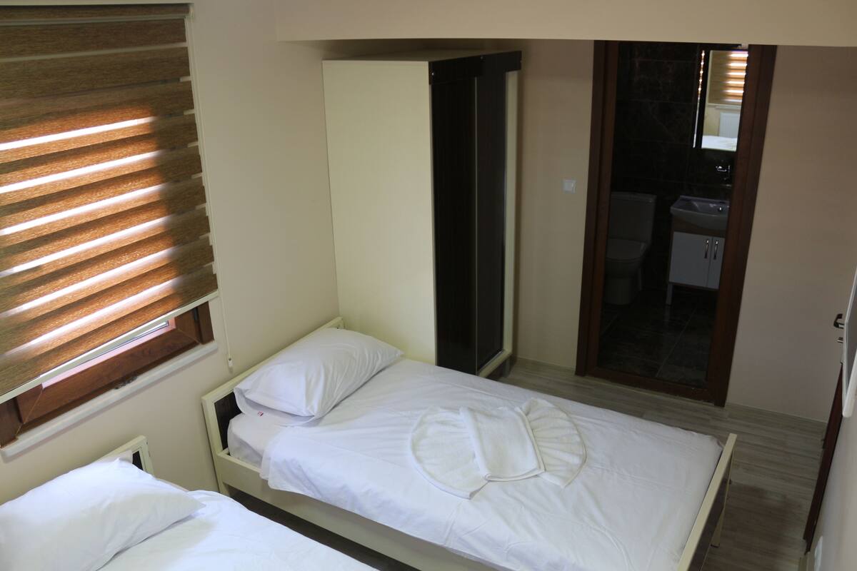 A small, single-bed room with a wooden headboard, a window with blinds, and a bathroom visible through an open door.