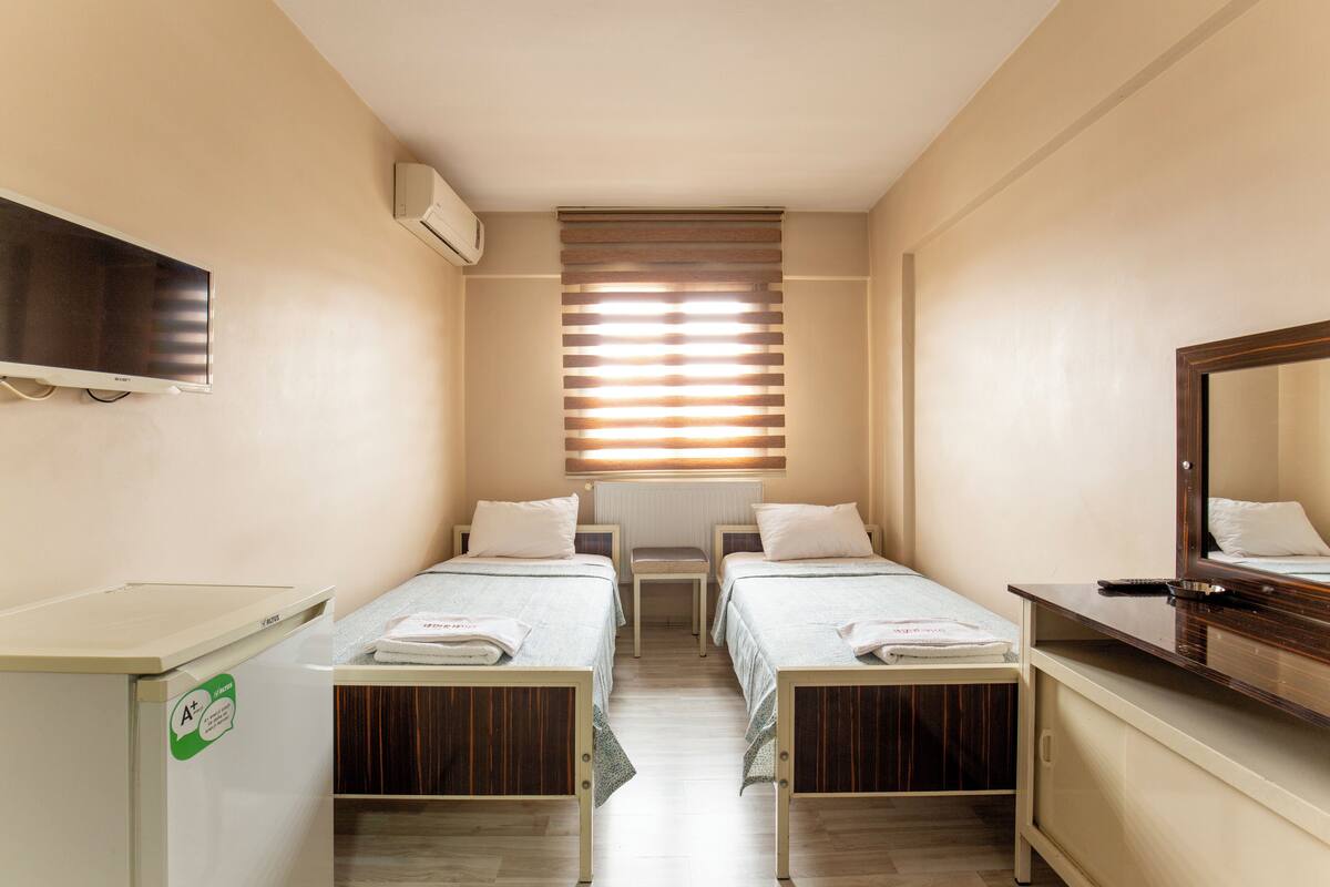 A small, well-lit room with two single beds, a desk, a chair, a mirror, and a television.