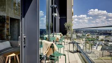 A modern balcony with green chairs and a sofa, a glass door, and a city view.