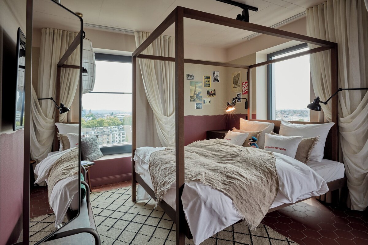 Un lit à baldaquin avec vue sur la ville, une table de chevet avec une lampe et un mur orné de diverses photos et affiches.