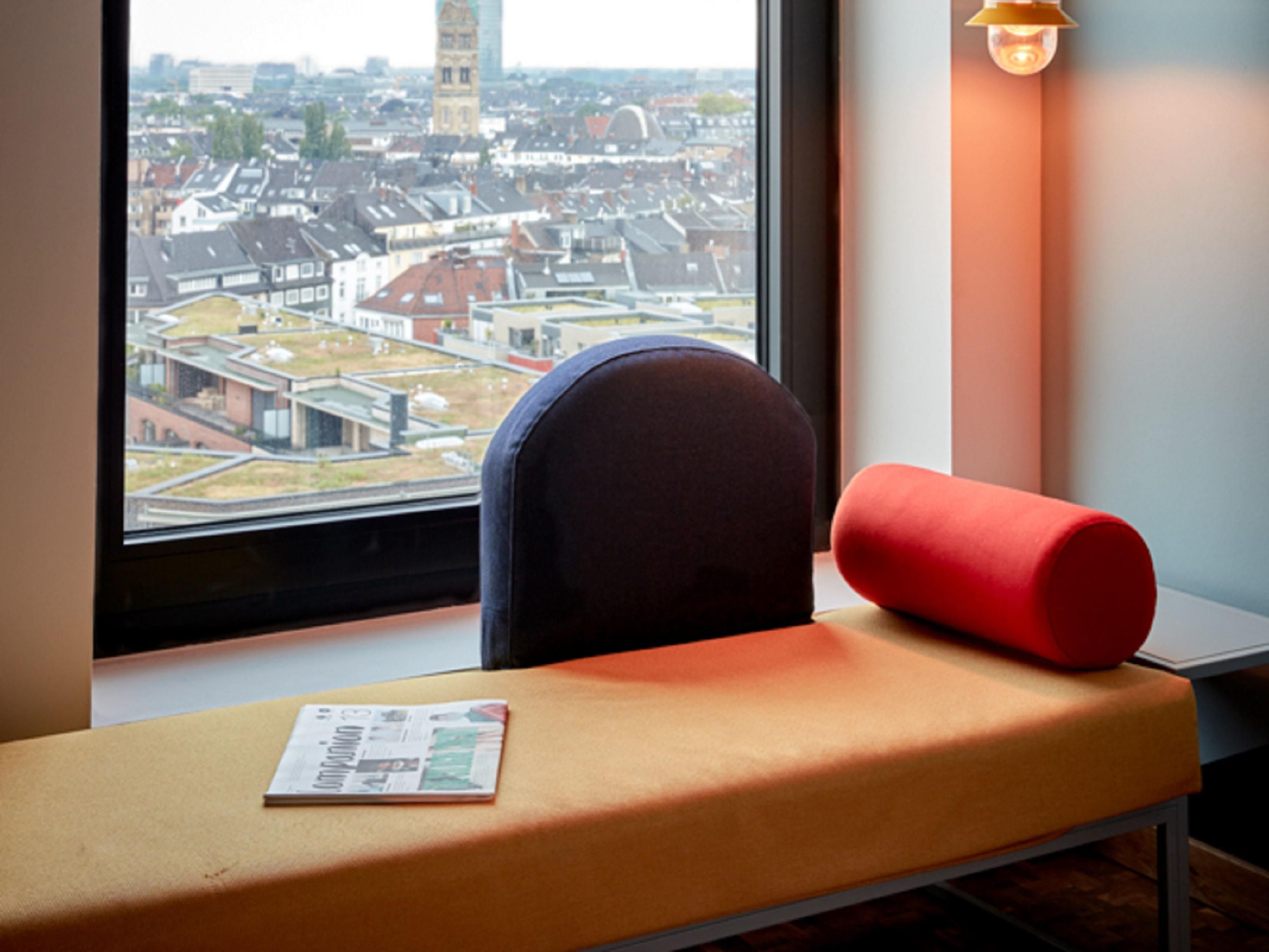 A window with a view of a cityscape, a chair with a cushion, and a lamp.