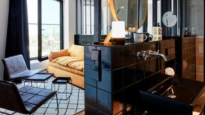 Una habitación de hotel moderna con un ventanal grande, un sofá, una mesa de centro, un espejo y un baño con pared de azulejos negros y un lavamanos.