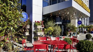 L’extérieur d’un hôtel moderne, avec un espace de repos en plein air, une enseigne indiquant « The Paris Club » et un panneau annonçant un service hôtelier disponible 24 heures sur 24.