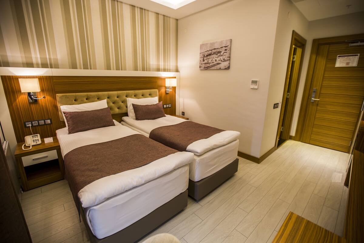 Habitación de hotel con dos camas, cabeceros de madera, lámparas de noche, un teléfono y un cuadro enmarcado en la pared.