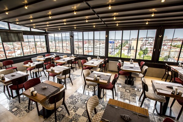 Un restaurant spacieux, doté de nombreuses tables et chaises, de grandes fenêtres offrant une vue sur la ville et d’un plafond au design moderne.