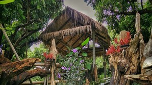 A rustic hut with a thatched roof surrounded by lush greenery and flowering plants. - Mon Saeng Jun Homestay (Phrao)