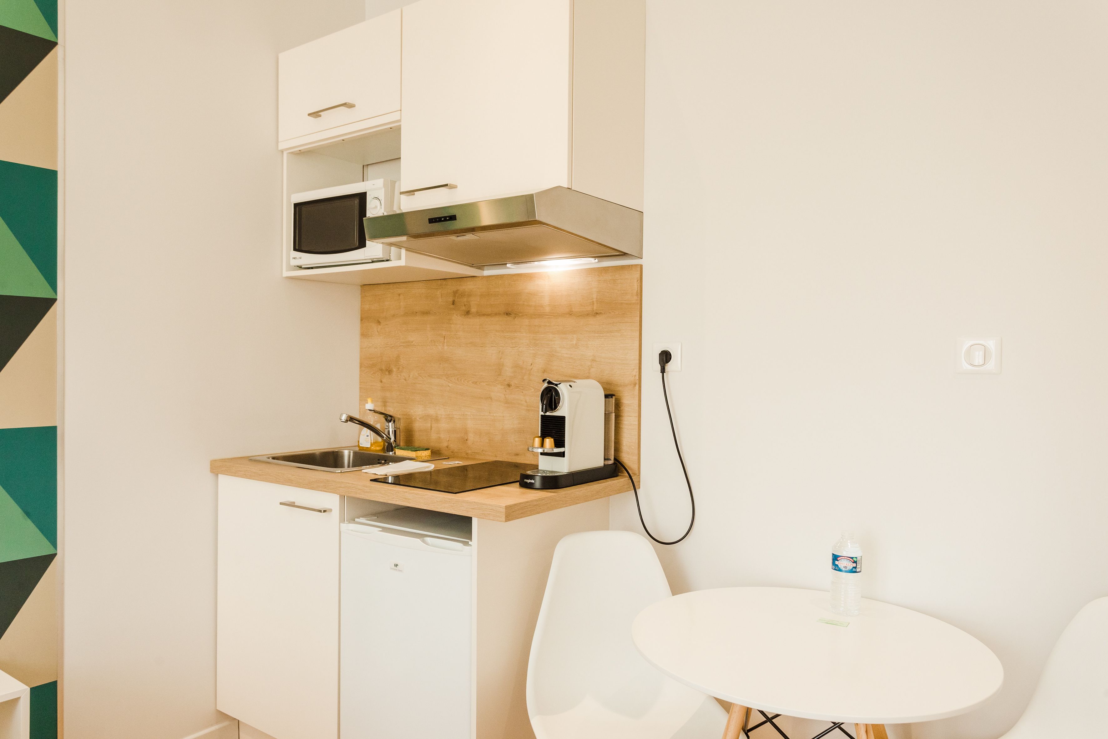 A compact kitchen with a microwave, coffee maker, and sink. There is a small table with a chair and a water bottle.