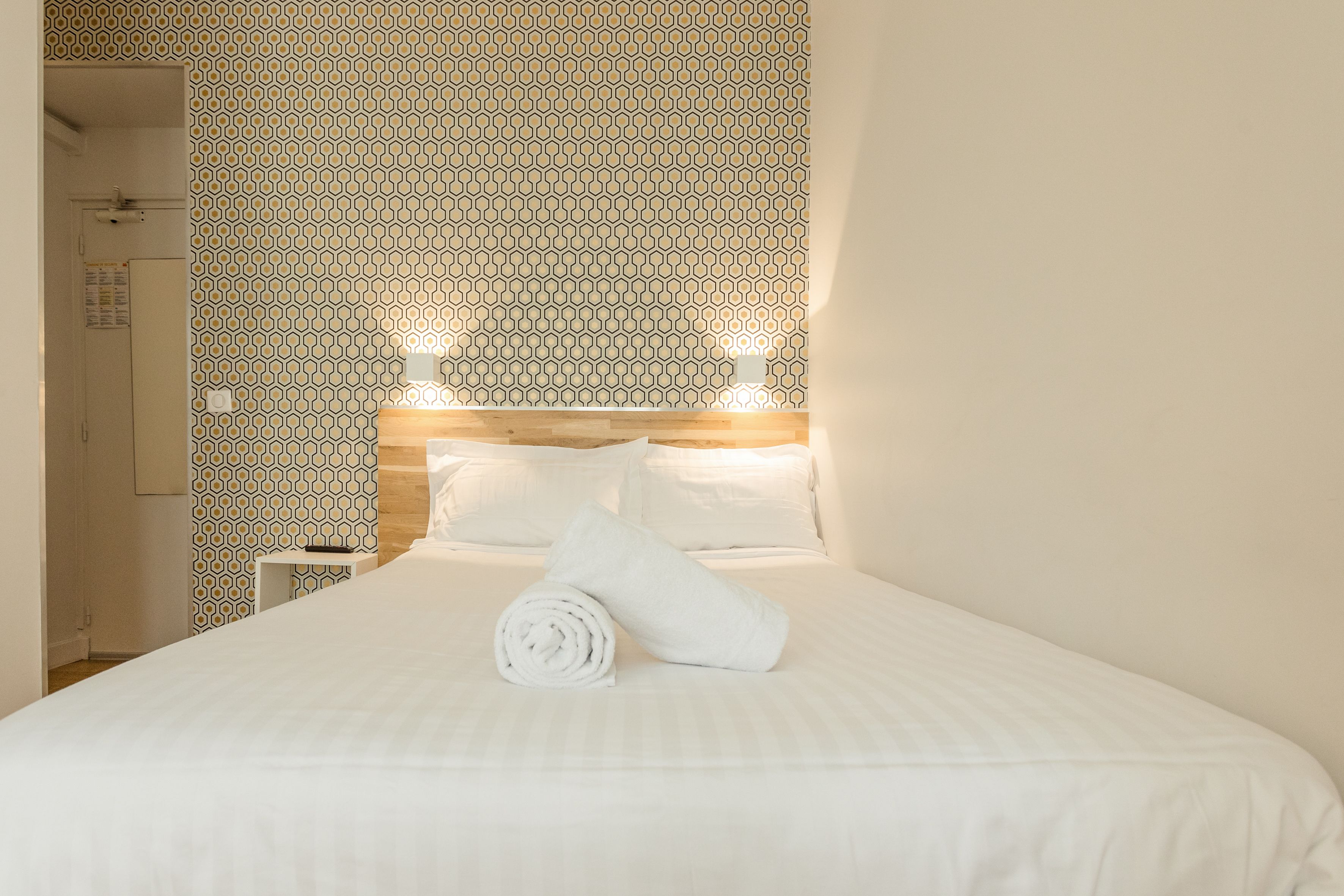 A neatly made bed with white linens, a wooden headboard, and a patterned wall.