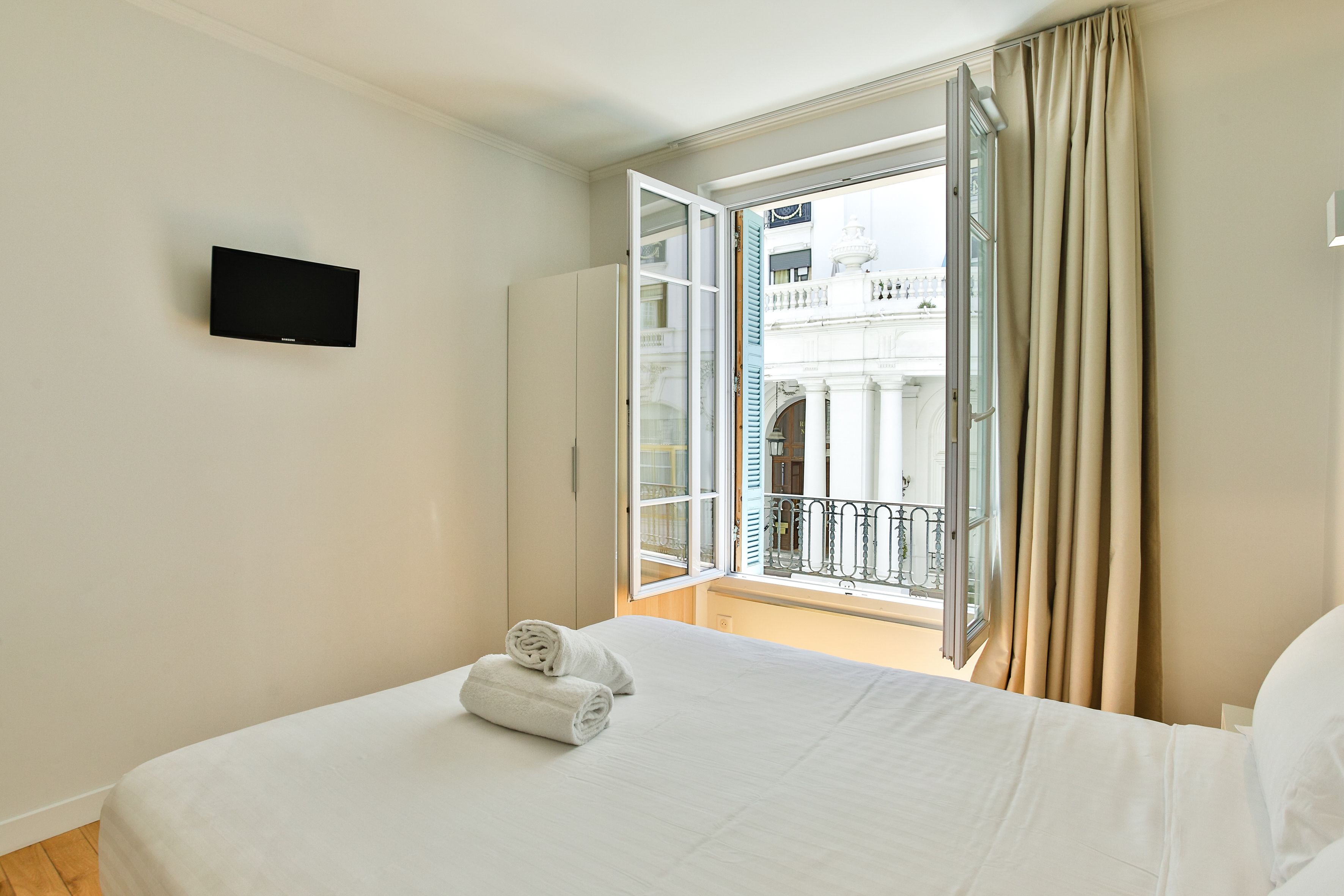 A hotel room with a bed, a flat-screen TV, and an open balcony with curtains.
