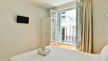 A hotel room with a bed, a flat-screen TV, and an open balcony with curtains.