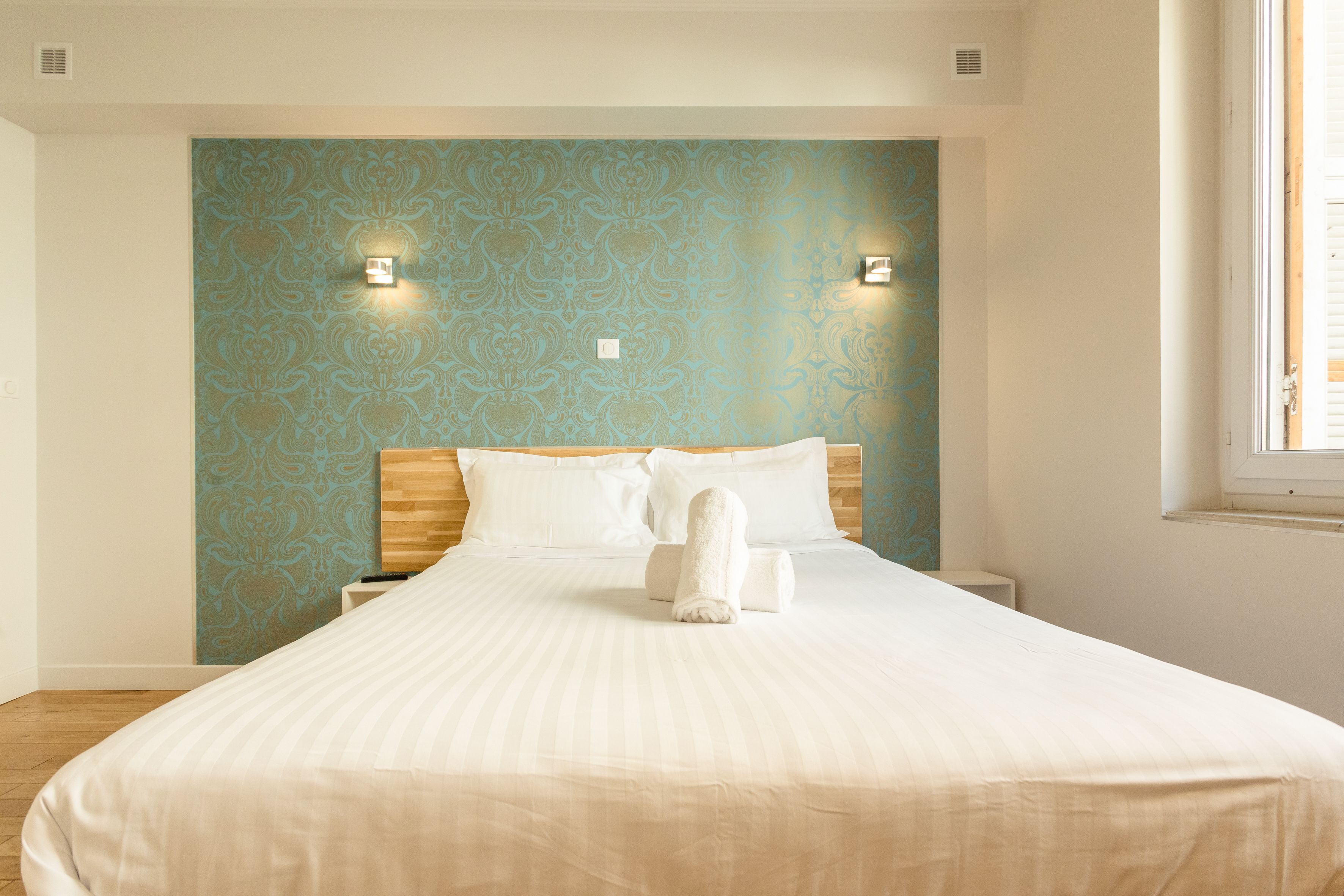 Een net opgemaakt bed met een witte deken en kussens, een houten hoofdbord en een muur met blauw behang met een patroon.