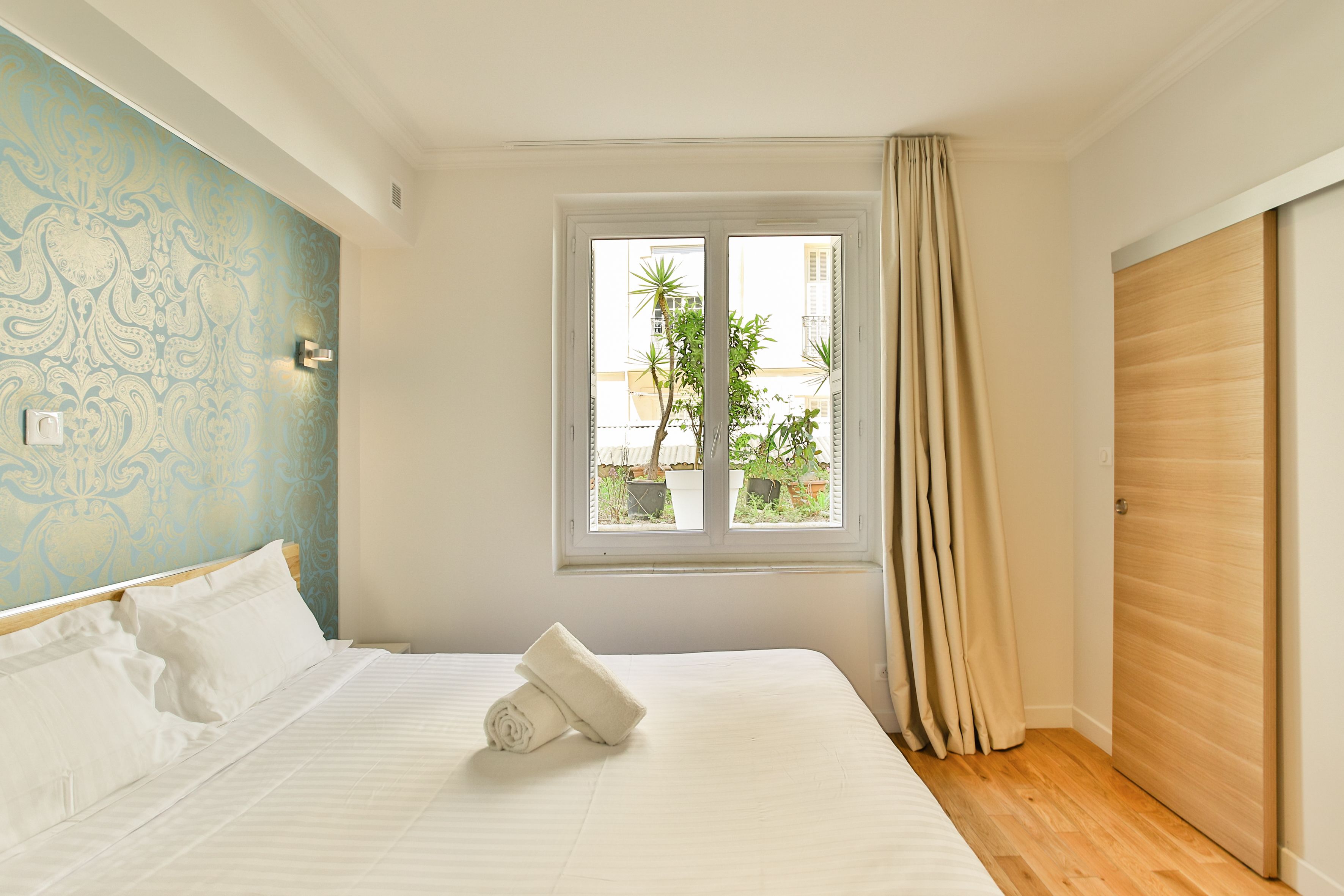 A neatly made bed with a rolled towel, a window with curtains, and a view of greenery outside.