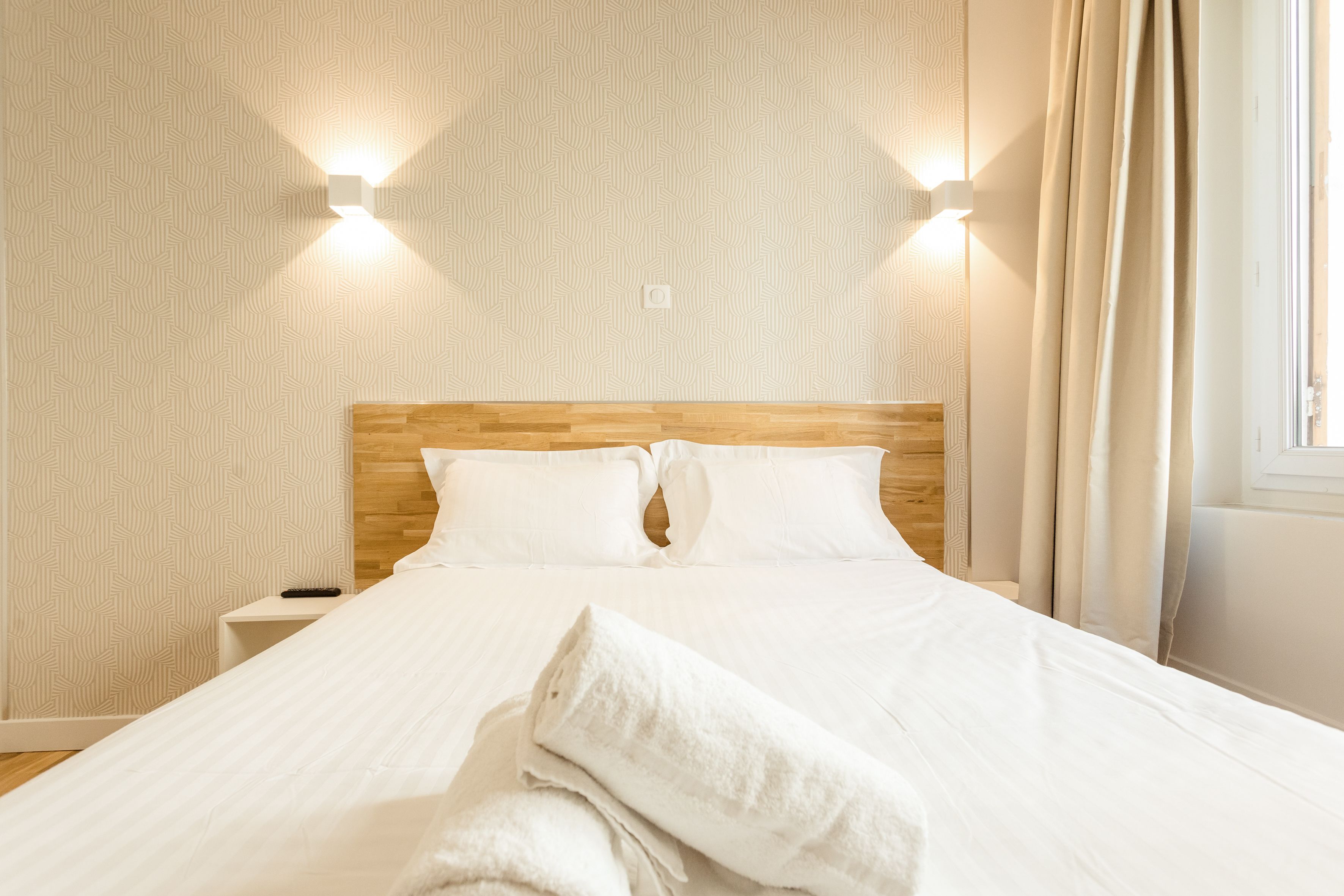 A neatly made bed with white linens, a wooden headboard, and two wall-mounted lights.