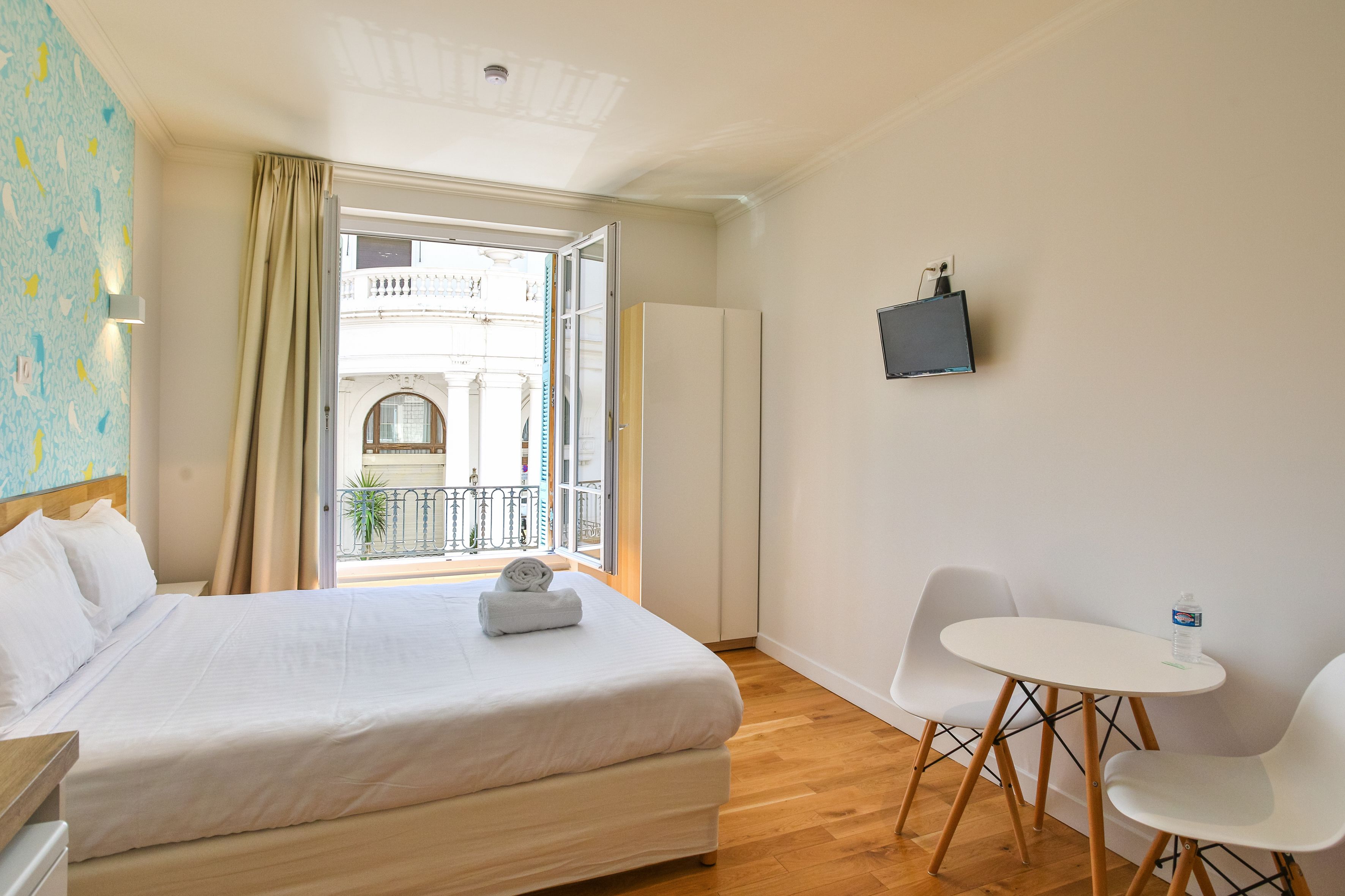 A hotel room with a bed, a small table with chairs, a TV mounted on the wall, and a balcony with a view of the city.