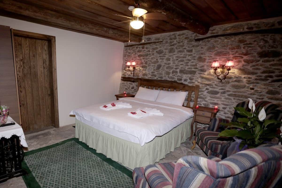 Une chambre confortable avec un lit en bois, des murs en pierre, un tapis à motifs verts et des fauteuils à rayures.