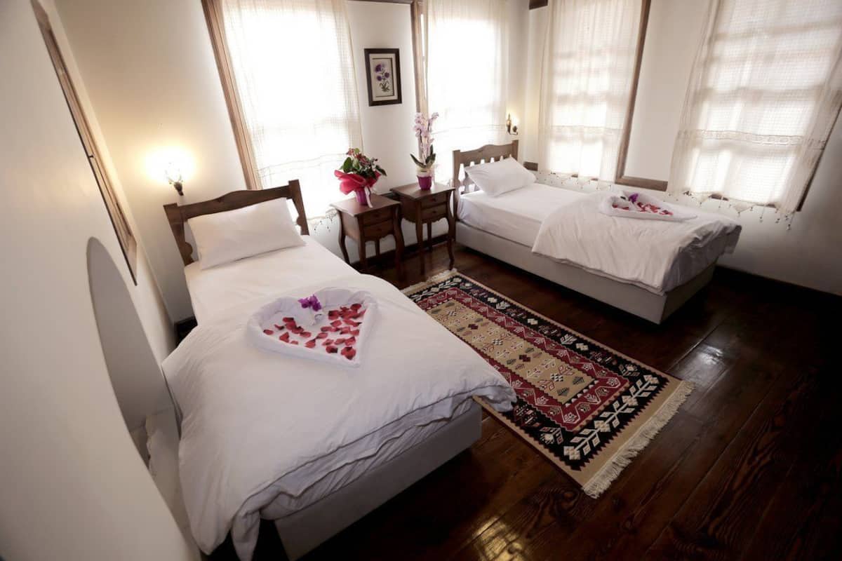 Une chambre d’hôtel avec deux lits simples, un bureau en bois, une chaise, une petite table ornée d’un vase de fleurs et un tapis à motifs.
