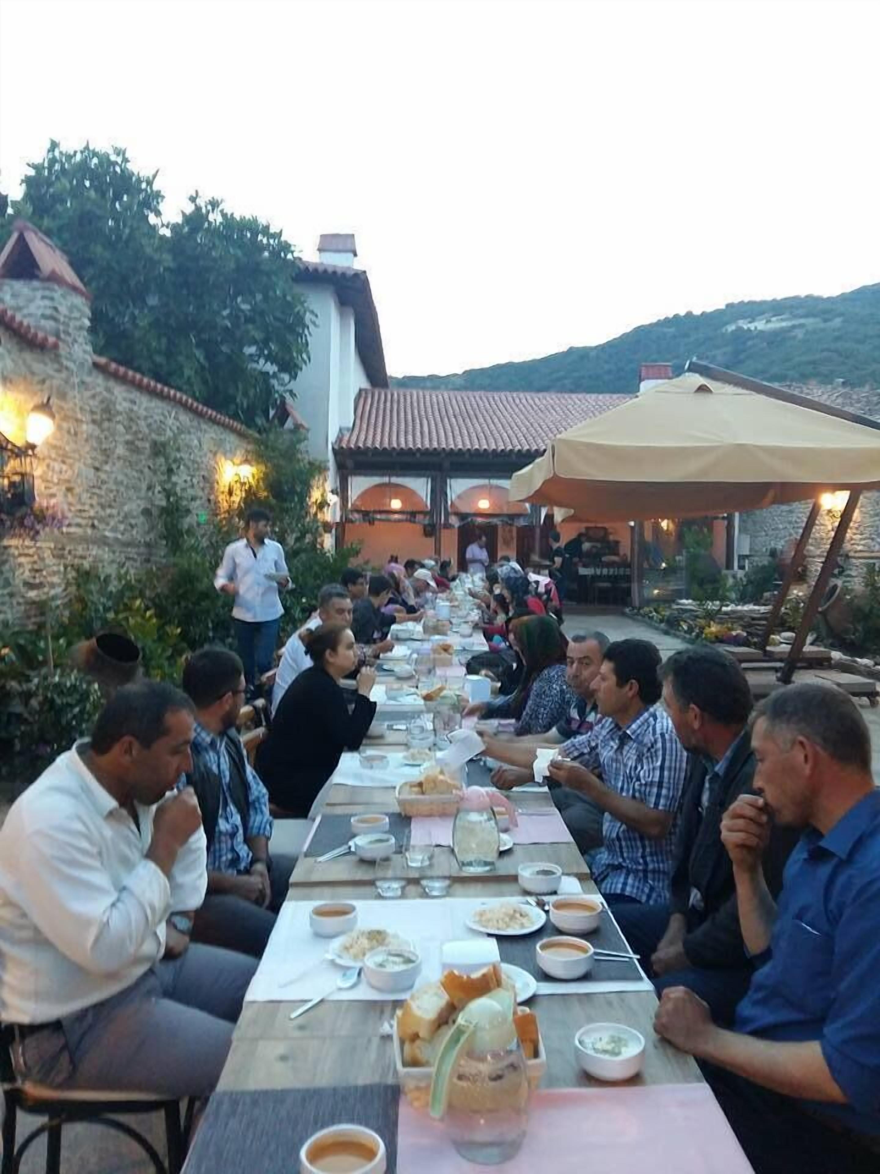 A long dining table with people seated on chairs, surrounded by a stone wall and a building with a tiled roof.