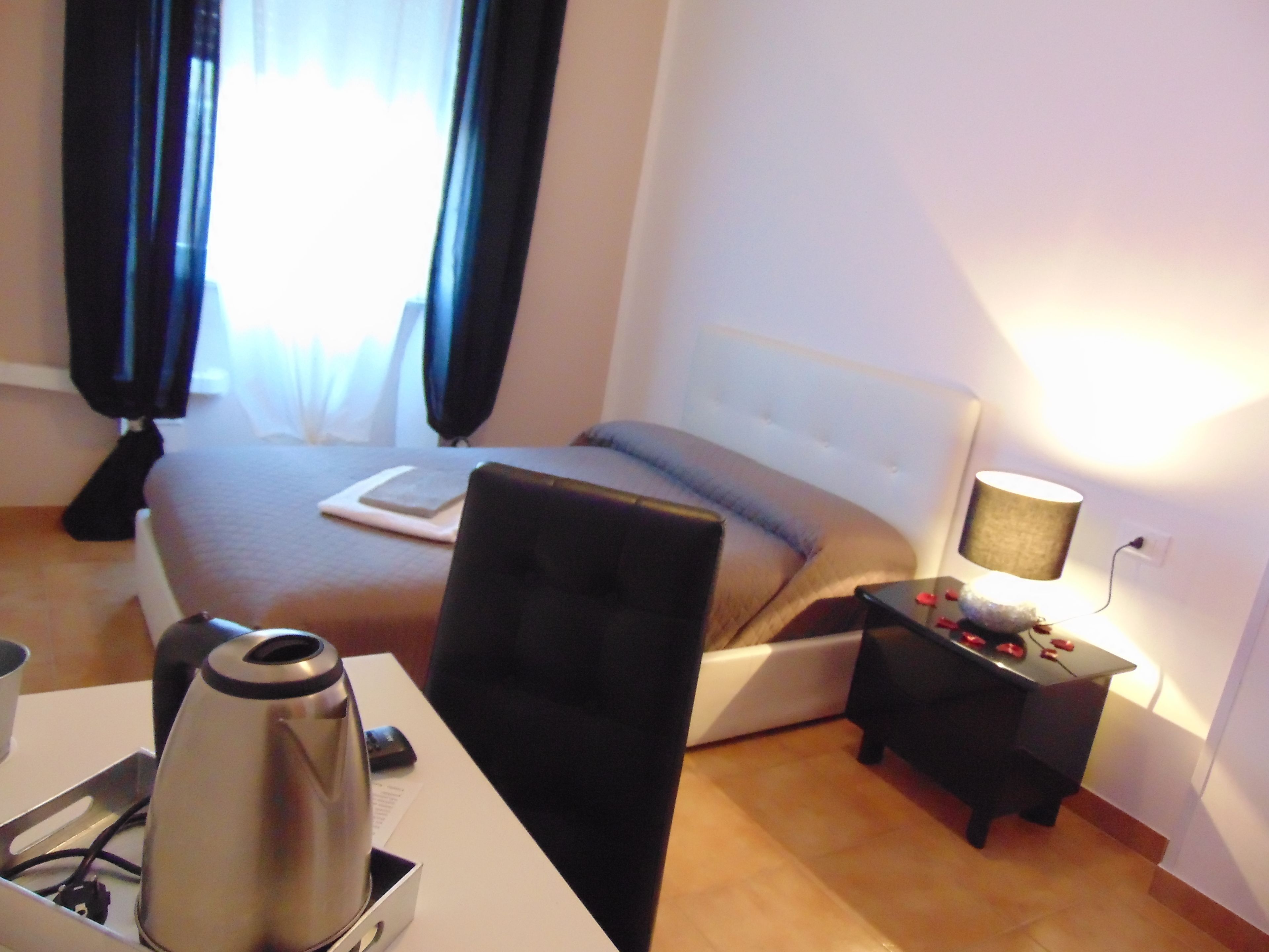 A hotel room with a bed, a desk with a kettle, a chair, a small table with a lamp, and a window with curtains.