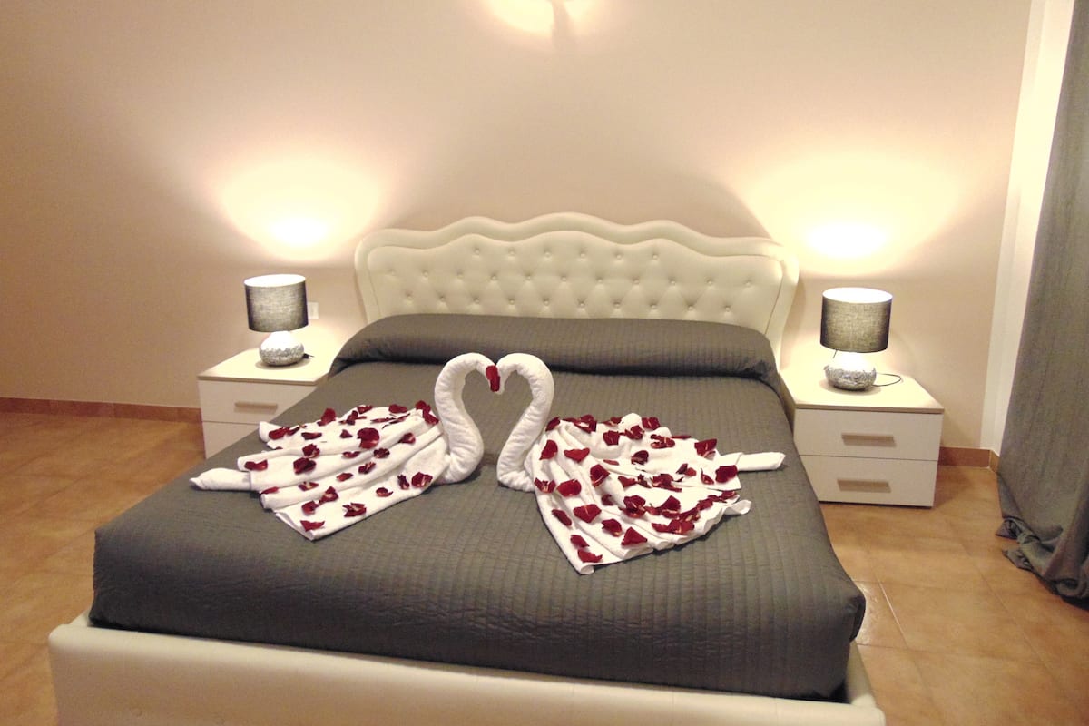 A neatly made bed with a grey bedspread and two white towels arranged in swan shapes with red rose petals. There are two bedside lamps on white nightstands.