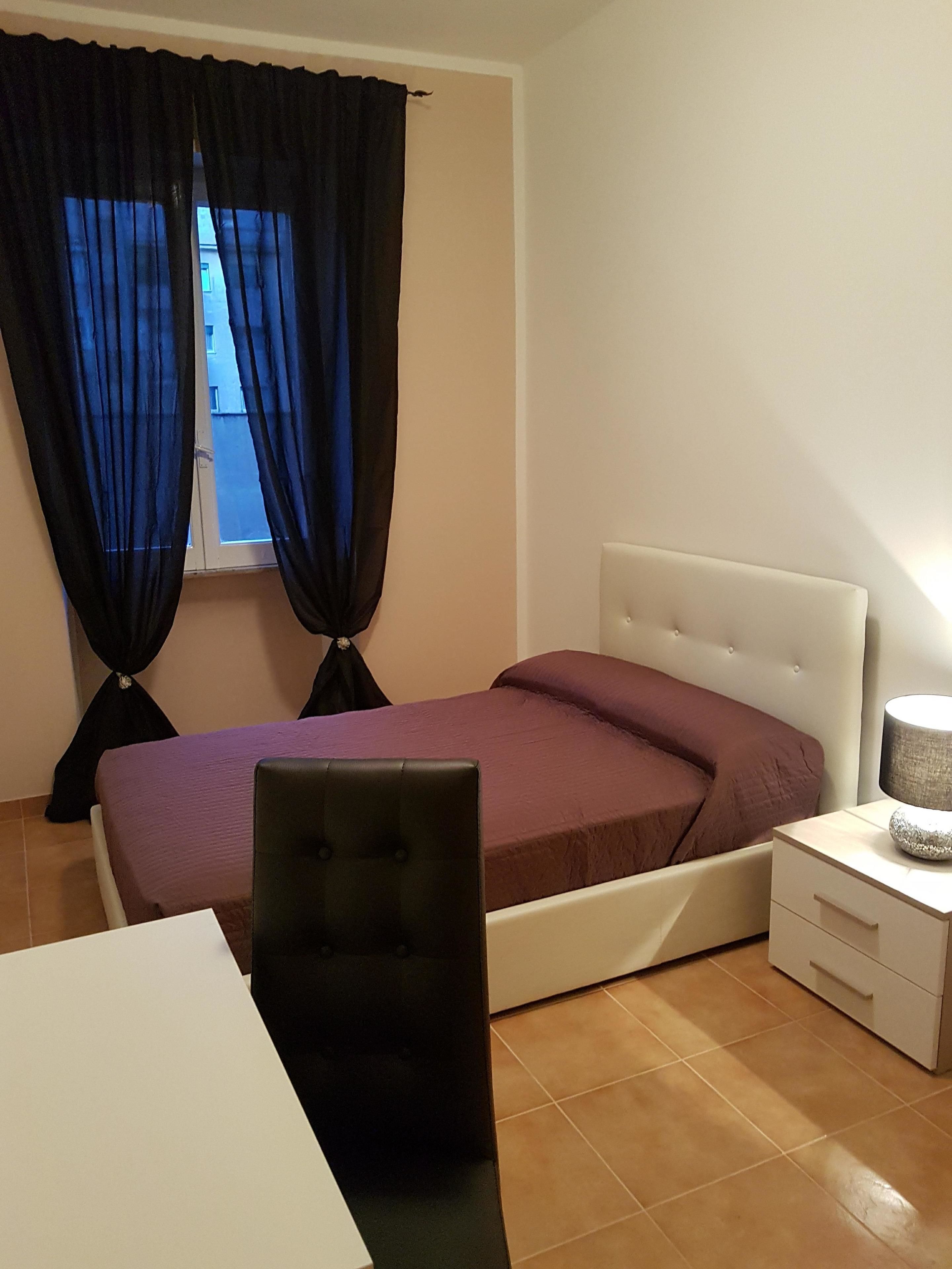 Ein Schlafzimmer mit Bett, Nachttisch, Sessel und einem Fenster mit Vorhängen.