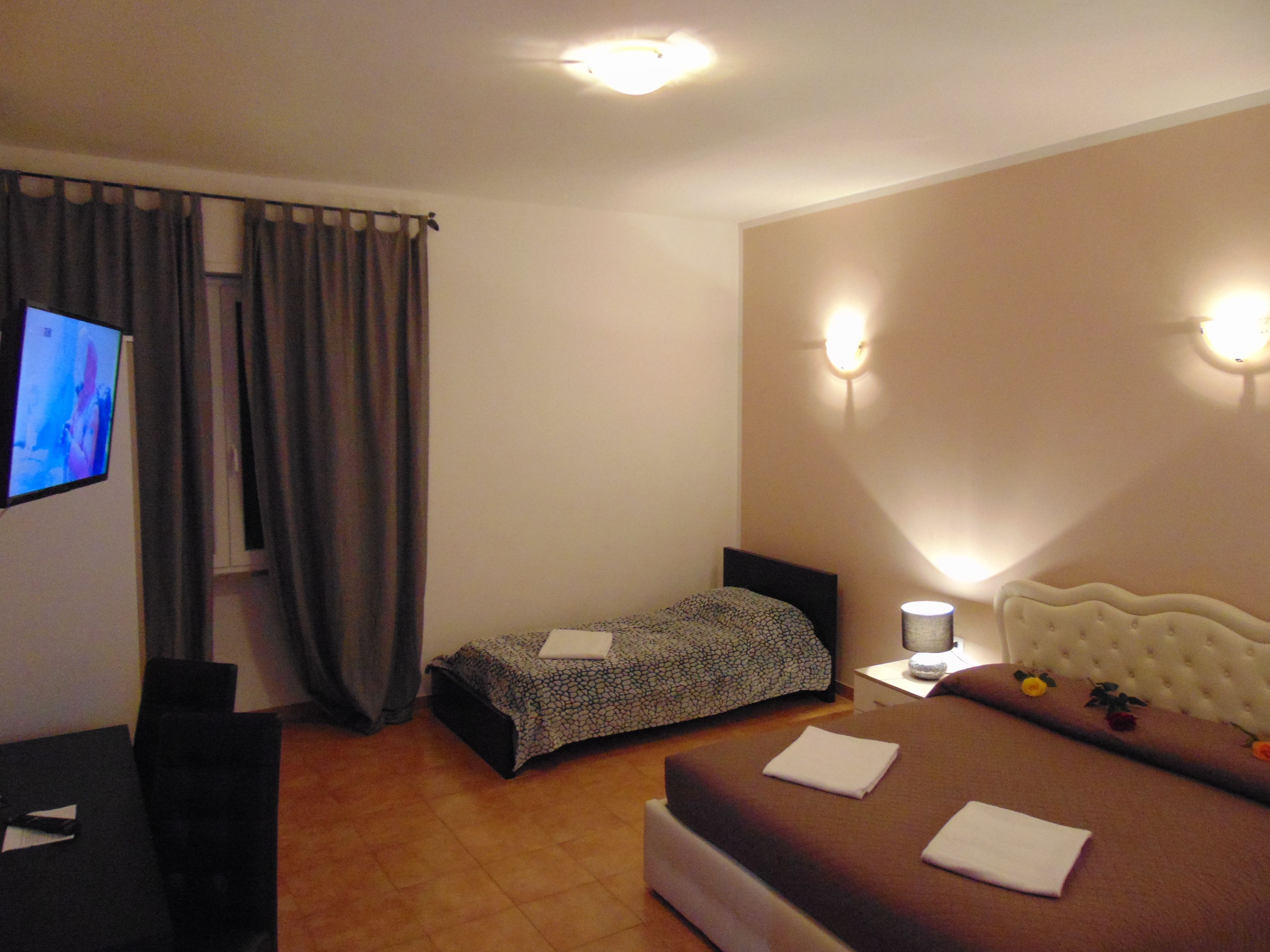 Habitación de hotel con cama, mesita de noche con lámpara, televisor en la pared y ventana con cortinas.
