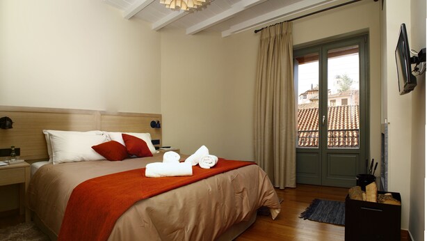 A hotel room with a bed, a nightstand, a window with curtains, and a view of buildings outside. - Kazas Luxury Apartments (Gortynia)