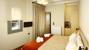 A hotel room with a bed, a window, a curtain, a door, a wall-mounted TV, and a bedside table. - Kazas Luxury Apartments (Gortynia)