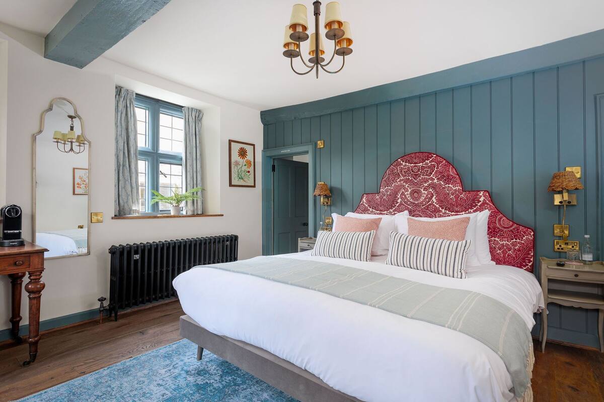 A bedroom with a large bed, a dresser, a radiator, a window with curtains, and a chandelier.