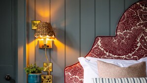 Ein ordentlich bezogenes Bett mit gemusterten Kissen, einer Nachttischlampe und einem kleinen Tisch mit Pflanze und Büchern.