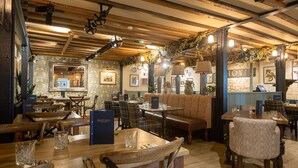 Ein gemütliches Restaurant-Interieur mit Holzbalken, gemustertem Wandpapier und verschiedenen Sitzmöglichkeiten, darunter Tische und Bänke.