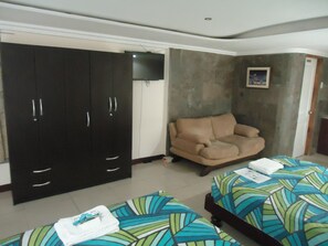 Quarto de hotel com guarda-roupa espaçoso, sofá, duas camas com roupa de cama colorida e uma mesinha de centro com um controle remoto.