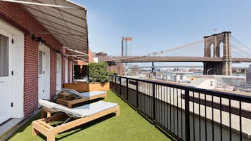 Terraço no telhado com duas cadeiras de descanso, uma mesa de madeira pequena e um vaso de planta. Um guarda-corpo de metal preto circunda o terraço, e uma grande ponte é visível ao fundo.