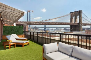 A rooftop terrace with two lounge chairs, a small table, and a sofa. A green hedge and a metal railing are visible. In the background, a large suspension bridge and a construction crane are seen. - 33 Hotel, New York City, Seaport (New York)