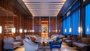 Een moderne lounge met een centrale ronde tafel, omringd door banken en fauteuils, een bar op de achtergrond en grote ramen met gordijnen.