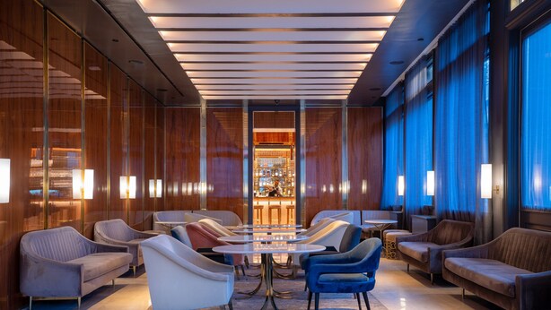Un área de estar moderna con una mesa redonda central rodeada de sofás y sillones, un bar al fondo y grandes ventanas con cortinas.
